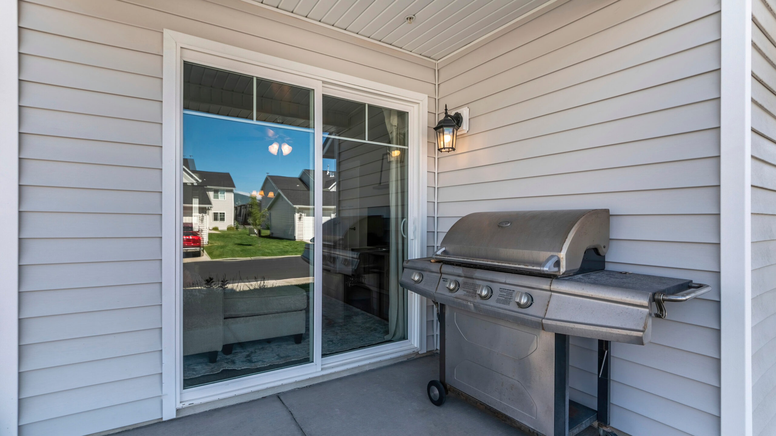 Pano Exterior of a house with a sliding glass door and vinyl sidings. There is a gas griller near the wall by the entrance and a wall lamp near the door.